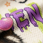 Close-up of a white knit fabric with a colorful graphic print featuring purple dripping letters, lime green accents, a pink circular element, and a black-and-white animal face.
