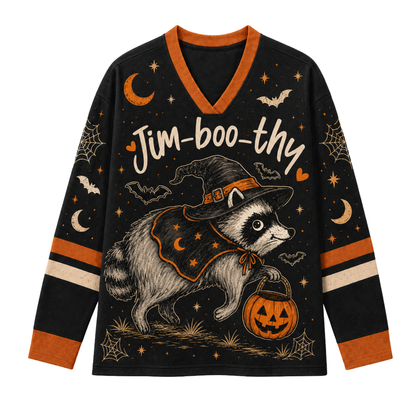 A long-sleeve black V-neck sweater with orange and beige accents, featuring a cartoon raccoon dressed as a witch holding a jack-o'-lantern, with the text 'Jim-boo-thy' and Halloween motifs like bats, moons, stars, and spiderwebs on a white background.