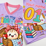 A collage showing a children's V-neck t-shirt with customizable printed graphics. The shirt features a light purple base with a pink collar, decorated with cute cartoon illustrations including a smiling dumpling in a basket, books, pencils, and the text 'Your name', 'in my KIND', and 'Marigold Ivy'.