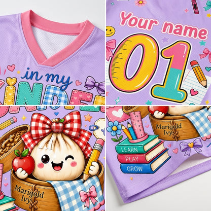 A collage showing a children's V-neck t-shirt with customizable printed graphics. The shirt features a light purple base with a pink collar, decorated with cute cartoon illustrations including a smiling dumpling in a basket, books, pencils, and the text 'Your name', 'in my KIND', and 'Marigold Ivy'.