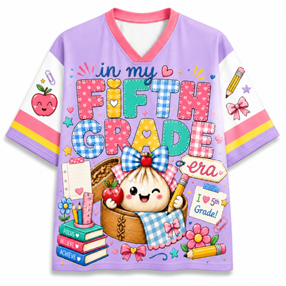 A purple V-neck t-shirt with a colorful cartoon graphic celebrating 'Fifth Grade'. The design features a cute dumpling character in a basket, books, apples, and school supplies, with the text 'in my FIFTH GRADE era'.