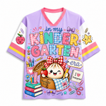A lavender short-sleeve V-neck t-shirt with a colorful cartoon graphic on the front reading 'in my KINDER GARTEN era'. The design features a cute girl with a red-and-white checkered bow, a basket, books, scissors, and other school-themed illustrations.