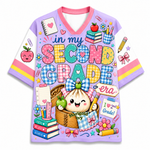 A short-sleeve V-neck t-shirt with a colorful school-themed graphic print. The design features the text 'in my SECOND GRADE era' in playful, multicolored bubble letters, surrounded by cute illustrations of a cartoon dumpling character, books, apples, and school supplies.