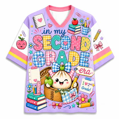 A short-sleeve V-neck t-shirt with a colorful school-themed graphic print. The design features the text 'in my SECOND GRADE era' in playful, multicolored bubble letters, surrounded by cute illustrations of a cartoon dumpling character, books, apples, and school supplies.