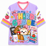 A short-sleeve V-neck t-shirt with a light purple body, white sleeves, and a colorful cartoon school-themed graphic on the front. The graphic features the text 'in my THIRD GRADE' with a cute dumpling character holding an apple and pencil, surrounded by school items like books, notebooks, and stars.