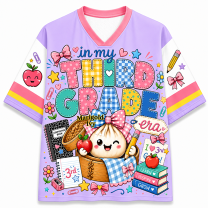 A short-sleeve V-neck t-shirt with a light purple body, white sleeves, and a colorful cartoon school-themed graphic on the front. The graphic features the text 'in my THIRD GRADE' with a cute dumpling character holding an apple and pencil, surrounded by school items like books, notebooks, and stars.