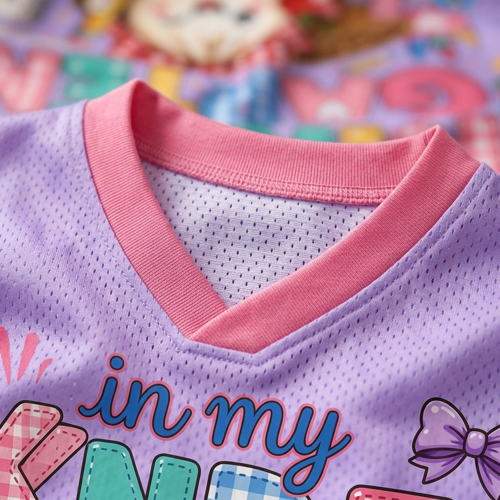 Close-up of a children's V-neck jersey top in light purple with a pink ribbed collar, featuring colorful graphic lettering and a bow illustration on the front.