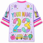 A light purple short-sleeve t-shirt with a colorful custom graphic design on the back. The design features the text 'YOUR NAME' in pastel letters, a large pastel '23' with smiley faces, crayons on the sleeves, and school-themed illustrations including an apple, bus, books, and globe at the bottom.