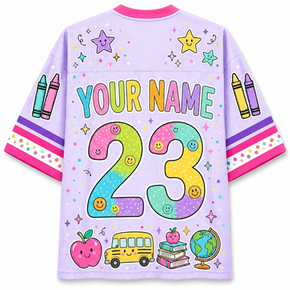 A light purple short-sleeve t-shirt with a colorful custom graphic design on the back. The design features the text 'YOUR NAME' in pastel letters, a large pastel '23' with smiley faces, crayons on the sleeves, and school-themed illustrations including an apple, bus, books, and globe at the bottom.