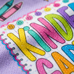 Close-up of a colorful children's notebook or journal cover with large glittery letters spelling 'KINDER', pastel crayon illustrations, smiley faces, and a purple textured background.