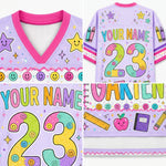 A personalized kids' V-neck jersey shirt shown in a four-panel collage, highlighting the front neckline, back with large customizable number 23 and name, front graphic detail, and back graphic detail. The shirt is light purple with hot pink trim and colorful school-themed graphics including smiley faces, stars, crayons, and a ruler.
