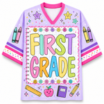 A light purple V-neck athletic-style jersey with a colorful 'FIRST GRADE' school-themed graphic on the front, including smiley faces, crayons, stars, a ruler, apple, notebook, and pencil illustrations.