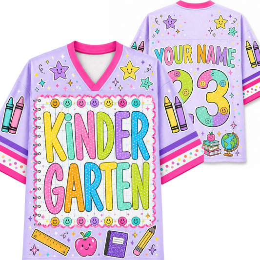A personalized children's V-neck t-shirt shown from both the front and back. The shirt is light purple with a pink collar and cuffs, decorated with colorful cartoon-style text reading 'KINDER GARTEN', stars, smiley faces, crayons, pencils, a ruler, an apple, and a notebook.
