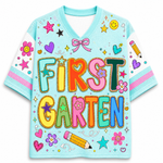 A light blue V-neck short-sleeve shirt with 'FIRST GARTEN' written in large, colorful, glittery letters. The shirt is decorated with various cartoon-style school-themed graphics including stars, hearts, flowers, bows, and a pencil on a white background.