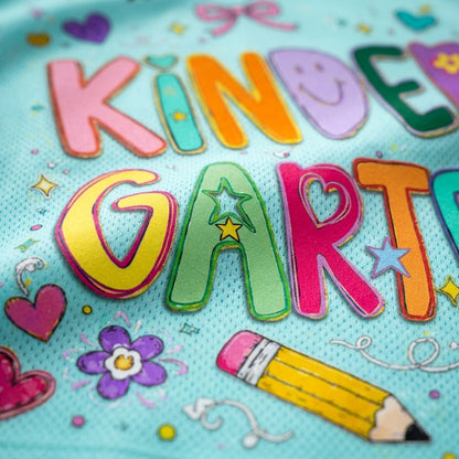 Close-up of colorful embroidered letters spelling 'KINDER GARTEN' on a light blue textured fabric, surrounded by small decorative motifs including flowers, stars, hearts, and a yellow pencil