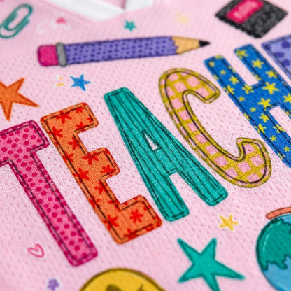 A close-up of a pink fabric surface featuring colorful embroidered or printed letters spelling "TEACH" in playful patterns, surrounded by school-themed motifs like stars, a pencil, and a chalkboard eraser.
