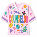 A pink short-sleeve V-neck t-shirt with a colorful school-themed graphic print featuring the word 'COUNSELOR' and illustrations of flowers, smiley faces, a globe, a backpack, and stationery on a white background.