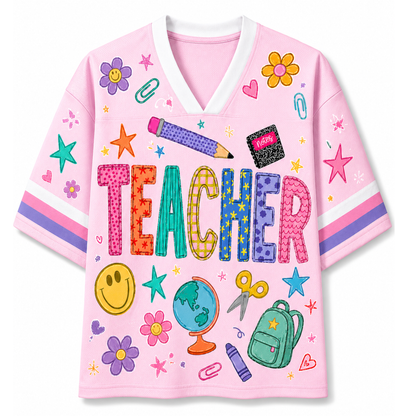 A pink short-sleeve V-neck T-shirt with a colorful school-themed graphic print featuring the word 'TEACHER' in patterned letters and illustrations of flowers, stars, paperclips, a globe, a backpack, and a smiley face.