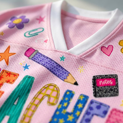 Close-up of a pink mesh V-neck jersey with colorful school-themed applique, including a purple pencil, flowers, paper clips, stars, hearts, and a notebook patch that says 'Notes'.