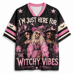 A women's short-sleeve V-neck jersey-style top with a black base and a large witch-themed graphic print. The design features three illustrated witches in pink and black attire, stars, bats, a crescent moon, and the text 'I'M JUST HERE FOR THE WITCHY VIBES.