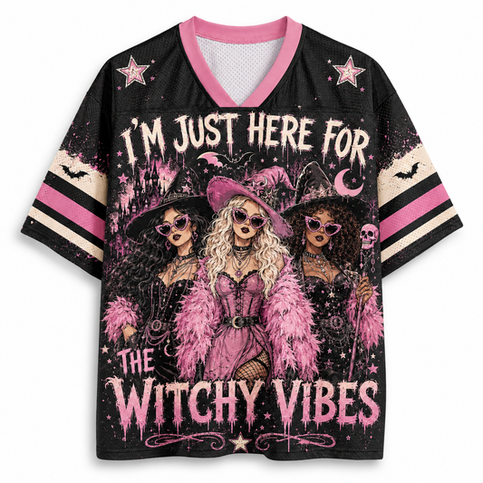 A women's short-sleeve V-neck jersey-style top with a black base and a large witch-themed graphic print. The design features three illustrated witches in pink and black attire, stars, bats, a crescent moon, and the text 'I'M JUST HERE FOR THE WITCHY VIBES.