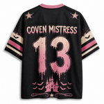 A black short-sleeve jersey-style shirt shown from the back, featuring pink and beige graphic details. The back has a pink collar, pink stars, the text 'COVEN MISTRESS' across the upper back, a large distressed pink number '13' in the center, and a pink castle with bats near the hem.