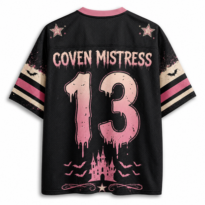 A black short-sleeve jersey-style shirt shown from the back, featuring pink and beige graphic details. The back has a pink collar, pink stars, the text 'COVEN MISTRESS' across the upper back, a large distressed pink number '13' in the center, and a pink castle with bats near the hem.