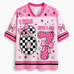 A pink short-sleeve V-neck t-shirt with a 'I WAS NOW I AM 6' birthday graphic. The number 6 is split into a black-and-white checkerboard pattern on the left and a pink leopard print pattern on the right, decorated with pink bows.
