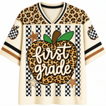 A short-sleeve V-neck t-shirt with a school-themed graphic. The shirt features a leopard print pattern on the shoulders and sleeves, a black and white checkered stripe design on the front, and a large central graphic of a leopard print apple with the words 'first grade' written in white script.