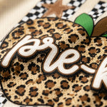 Close-up of a leopard-print patch with the word 'pre-k' in white script, featuring a green leaf detail and a black-and-white checkered background.