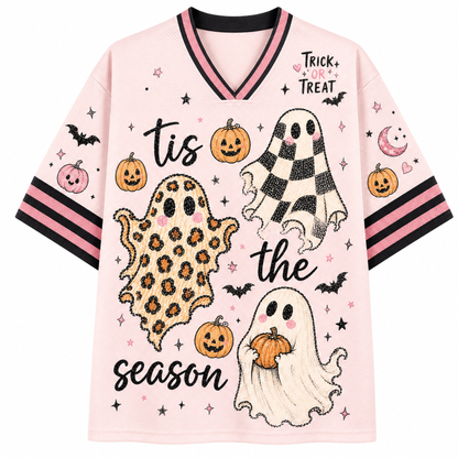 A light pink short-sleeve V-neck T-shirt with Halloween graphics, including three ghost illustrations (one leopard print, one black and white checkered, one plain white), pumpkins, bats, stars, and the text 'tis the season' and 'TRICK or TREAT'.