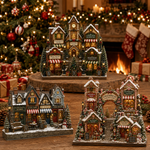 Three decorative Christmas village townhouse figurines displayed on a wooden surface in a festive holiday setting with a lit Christmas tree, fireplace, and stockings in the background.