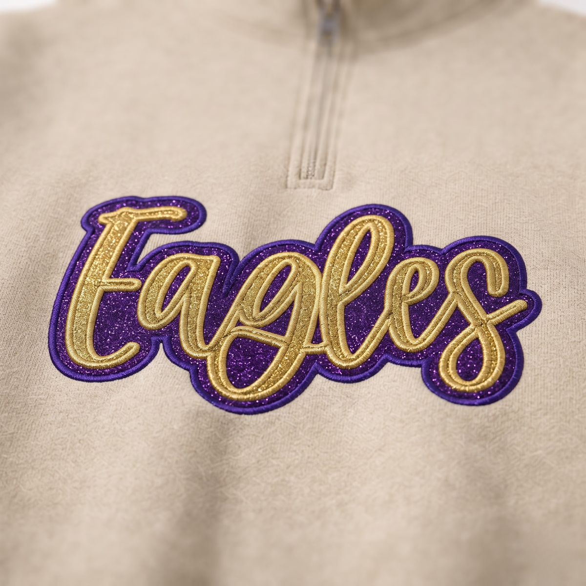 Close up shot of custom embroidered gold glitter Eagles cursive text with vibrant purple border on beige quarter-zip sweatshirt.