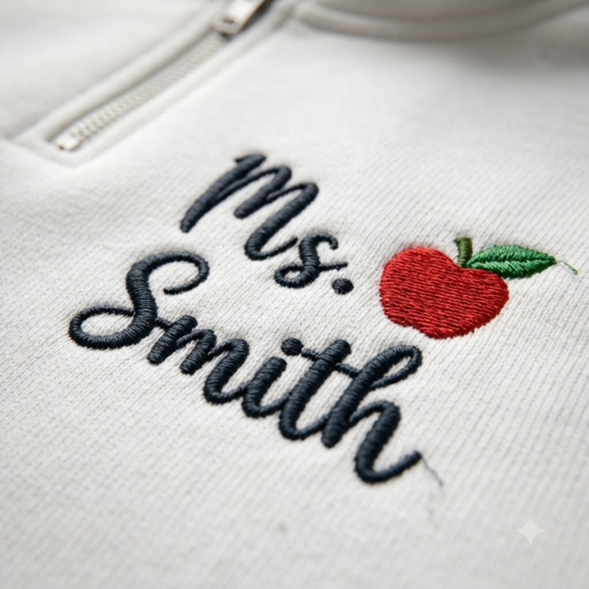 Close-up of a white zippered garment with embroidered text reading 'Ms. Smith' in dark navy script and a red apple with green leaves on the chest.