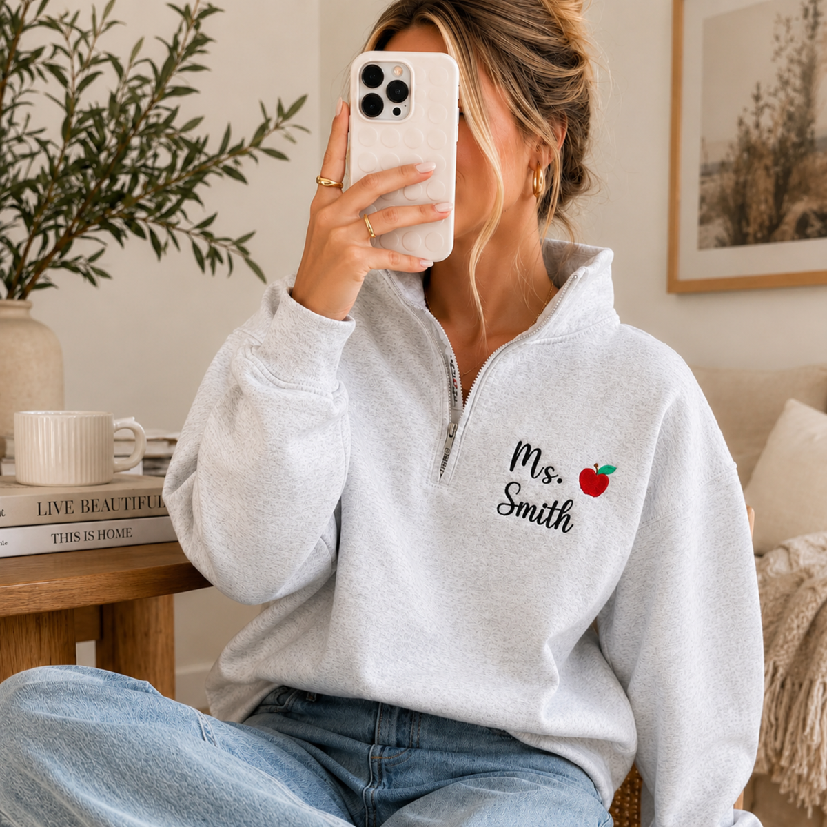 A woman takes a mirror selfie while wearing a light gray heathered quarter-zip sweatshirt with 'Ms. Smith' and a small red apple graphic embroidered on the left chest.