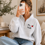 A woman takes a mirror selfie while wearing a light gray heathered quarter-zip sweatshirt with 'Ms. Smith' and a small red apple graphic embroidered on the left chest.