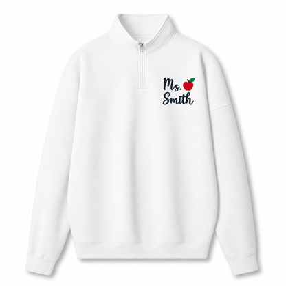 A white long-sleeve quarter-zip pullover sweatshirt with a mock neck collar, featuring the text 'Ms. Smith' in black script next to a small red apple graphic on the left chest.