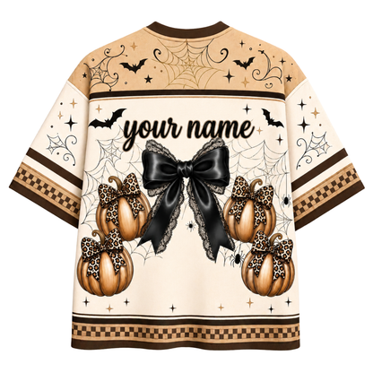 A Halloween-themed short-sleeve shirt shown from the back, featuring a cream base with brown and black spiderwebs, bats, stars, a large black bow, and three orange pumpkins with leopard-print bows. The center reads 'your name' in black script, indicating a customizable print.