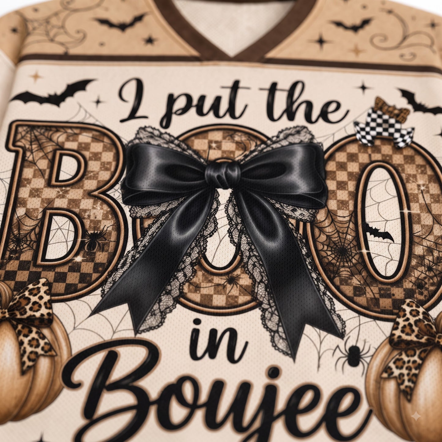 Close-up of a Halloween-themed graphic print featuring the phrase 'I put the BOO in Boujee' in black script over large decorative letters with a checkerboard pattern, a large black bow with lace trim, spiders, bats, and pumpkins on a cream background.