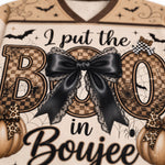 Close-up of a Halloween-themed graphic print featuring the phrase 'I put the BOO in Boujee' in black script over large decorative letters with a checkerboard pattern, a large black bow with lace trim, spiders, bats, and pumpkins on a cream background.