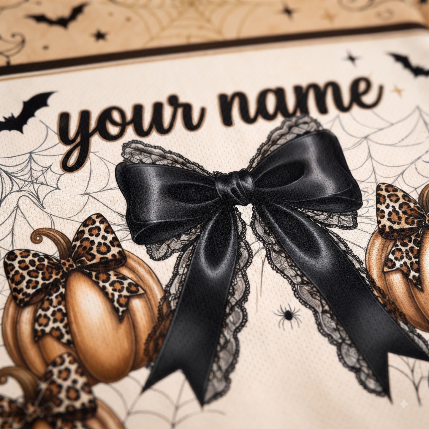 Close-up of a Halloween-themed personalized banner or print featuring the text 'your name' in black script, a large black satin bow with lace trim, illustrated pumpkins, spiderwebs, bats, and leopard-print accents on a cream background.
