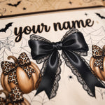 Close-up of a Halloween-themed personalized banner or print featuring the text 'your name' in black script, a large black satin bow with lace trim, illustrated pumpkins, spiderwebs, bats, and leopard-print accents on a cream background.