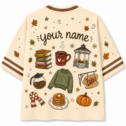 A cream-colored short-sleeve t-shirt with brown trim on the collar and cuffs, featuring a detailed autumn-themed graphic print with the text 'your name' and illustrations of a mug, sweater, pancakes, pumpkin, and books.