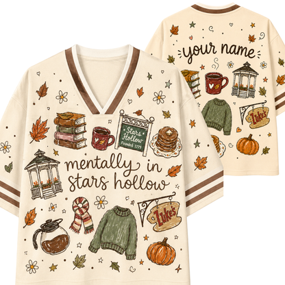 A cream-colored V-neck t-shirt with brown trim, featuring autumn-themed illustrations and the text 'mentally in stars hollow' on the front. The back view shows customizable text reading 'your name' along with additional seasonal graphics.