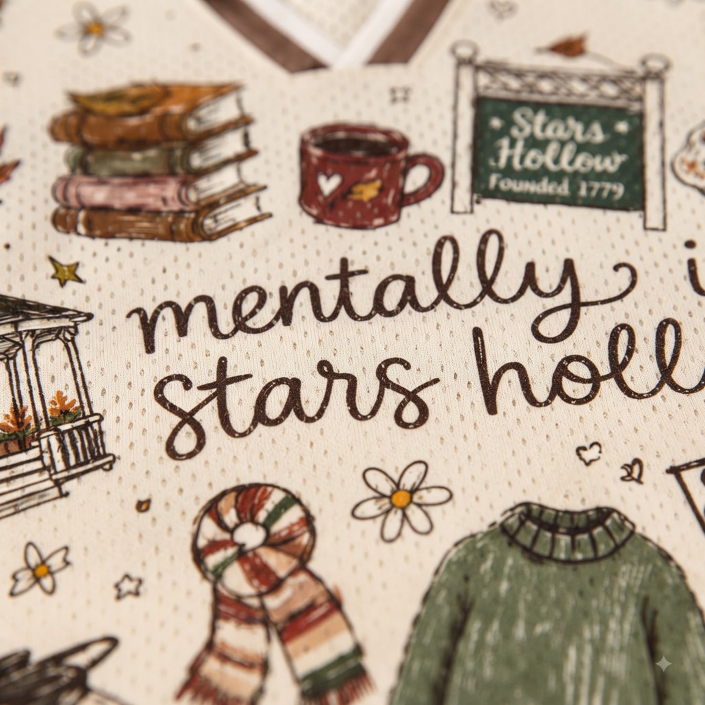 Close-up of a cream-colored fabric featuring a detailed winter-themed illustration and text that reads 'mentally in stars hollie'. The design includes drawings of a stack of books, a mug, a sign reading 'Stars Hollow Founded 1779', a porch, a striped scarf, and a green sweater.