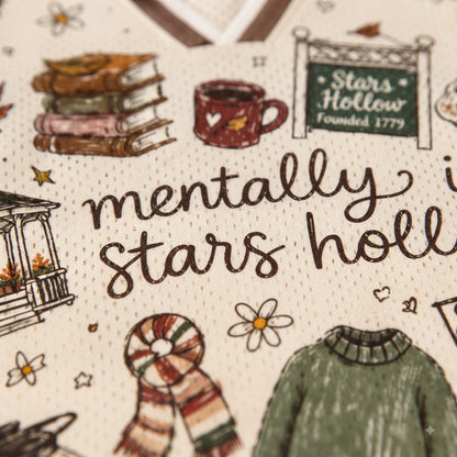 Close-up of a cream-colored fabric featuring a detailed winter-themed illustration and text that reads 'mentally in stars hollie'. The design includes drawings of a stack of books, a mug, a sign reading 'Stars Hollow Founded 1779', a porch, a striped scarf, and a green sweater.
