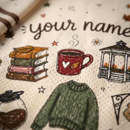 Close-up of a personalized fabric product, likely a towel or blanket, featuring a cream-colored woven background with a custom name printed in dark brown script. The fabric is decorated with various autumn-themed illustrations, including a stack of books, a red coffee mug with a white heart, a green sweater, a gazebo, and a coffee jar.