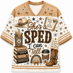 A short-sleeve V-neck t-shirt with a western-themed graphic print. The design features the text 'She's a SPED I can Tell' in a leopard-print patterned font, surrounded by illustrations of a cowboy hat, cowboy boots, books, a coffee cup, flowers, and stars in a brown, beige, and white color palette.