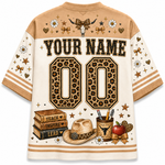 A short-sleeve custom sports jersey shirt shown from the back, featuring a tan and cream color scheme with leopard-print details, western-themed graphics including a cowboy hat, books, an apple, and a skull, and placeholder text 'YOUR NAME' and '00'.