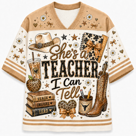 A short-sleeve V-neck t-shirt with a western-themed graphic print. The design features the text 'She's a TEACHER, I Can Tell' surrounded by illustrations of a cowboy hat, cowboy boots, a leopard-print notebook, books, an apple, and small hearts and stars.